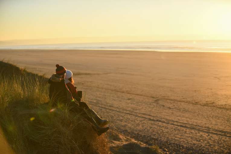 A couple sitting on a sand dune looking across a winter beach