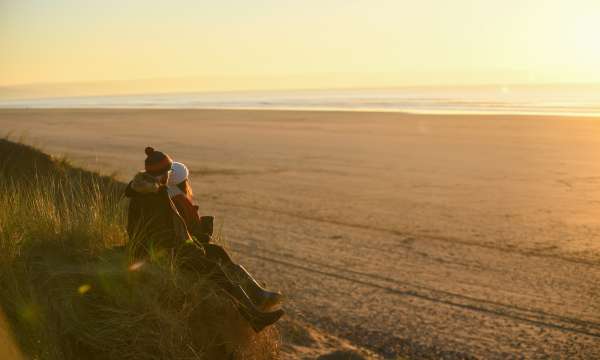 A couple sitting on a sand dune looking across a winter beach